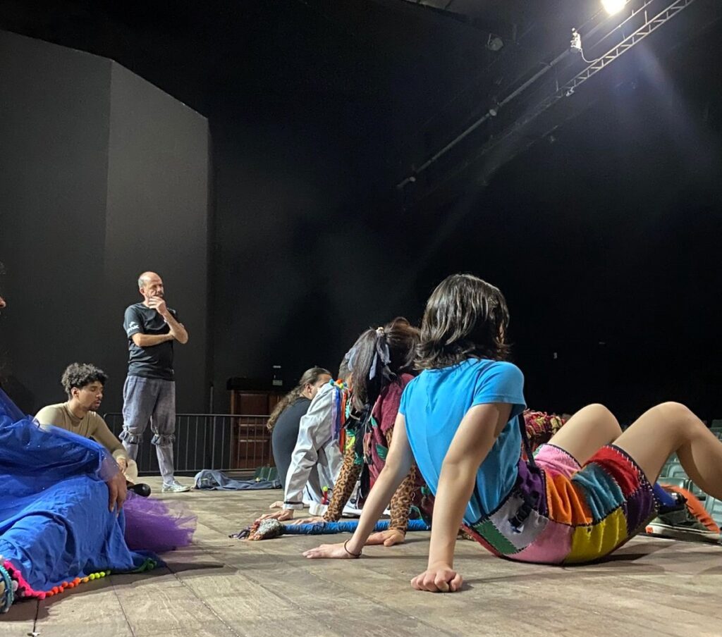 Elenco da Cia. Ceta sentado no palco durante ensaio, em processo de criação, com figurinos coloridos.