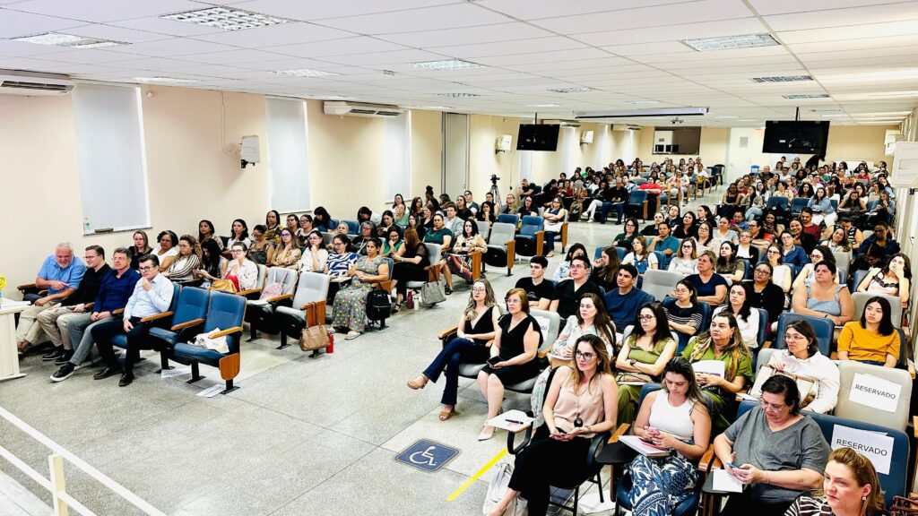 A imagem mostra uma sala de conferências lotada, com uma grande audiência sentada em cadeiras em fileiras. As pessoas estão vestidas com roupas casuais e formais, e a maioria parece estar prestando atenção em algo à frente, que não está visível na imagem. Há uma câmera em um tripé no fundo, sugerindo que o evento está sendo gravado ou transmitido. A iluminação é suave, vindo de cima, e as paredes são de cor clara. Há alguns equipamentos de projeção no teto. A sala parece ser um local de aprendizado ou apresentação, como uma universidade ou centro de treinamento.