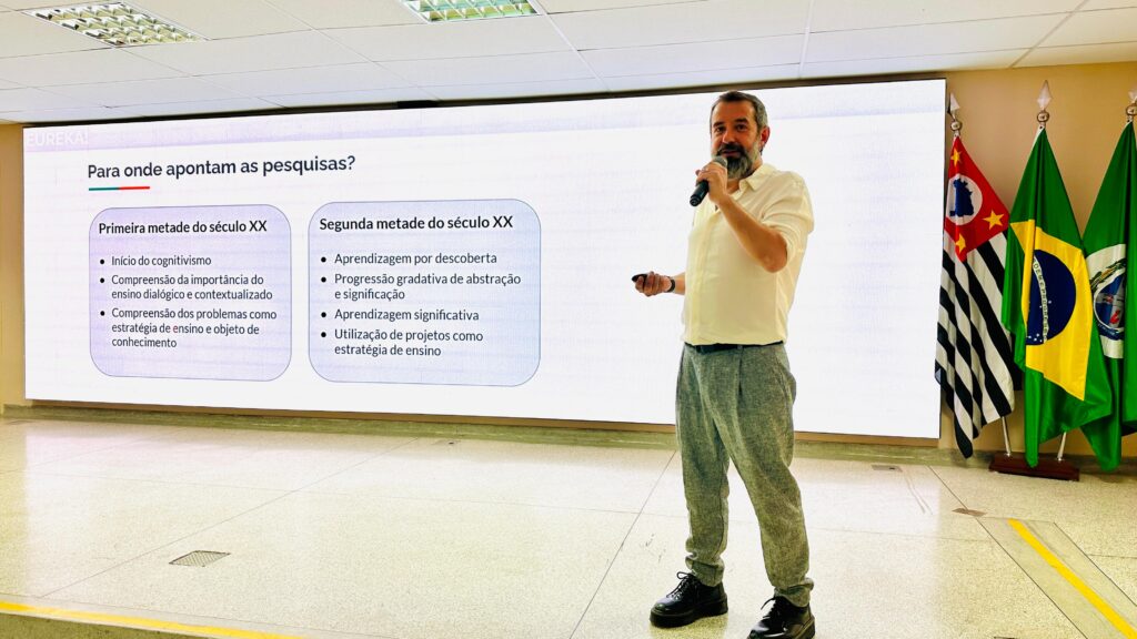 Um homem está em pé, falando ao microfone, em frente a uma tela de apresentação. A tela mostra um slide com texto em português sobre tendências de pesquisa na educação ao longo do século XX. O ambiente está bem iluminado e há quatro bandeiras ao fundo, incluindo a do Brasil e a do estado de São Paulo, indicando que o evento acontece em território brasileiro.