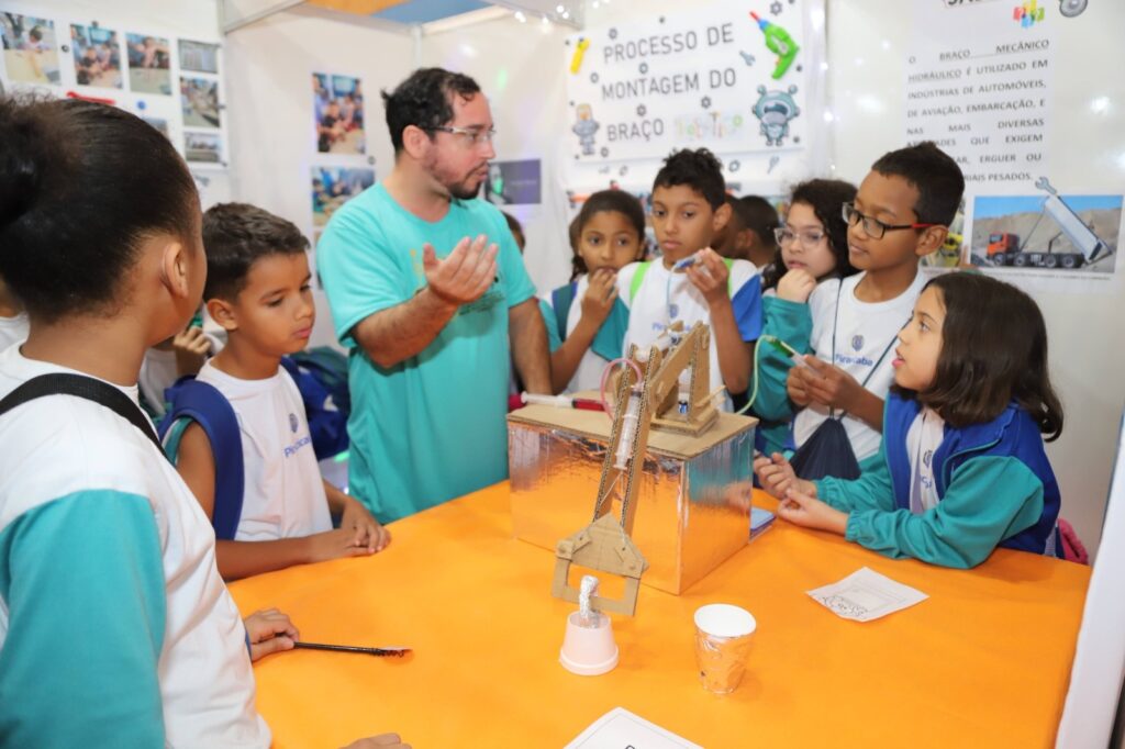 A imagem mostra um grupo de crianças em uma feira de ciências, observando atentamente um braço mecânico hidráulico em uma mesa. Um professor ou instrutor, vestindo uma camiseta verde, está explicando o funcionamento do projeto para as crianças. O braço mecânico, feito de papelão e com seringas para o sistema hidráulico, está montado sobre uma caixa coberta com papel alumínio. Ao fundo, há um painel com informações sobre o projeto, incluindo o título "PROCESSO DE MONTAGEM DO BRAÇO MECÂNICO HIDRÁULICO" e imagens relacionadas a indústrias e veículos pesados. As crianças, algumas usando uniformes escolares com o nome "Piracicaba", demonstram interesse e curiosidade enquanto interagem com o projeto.