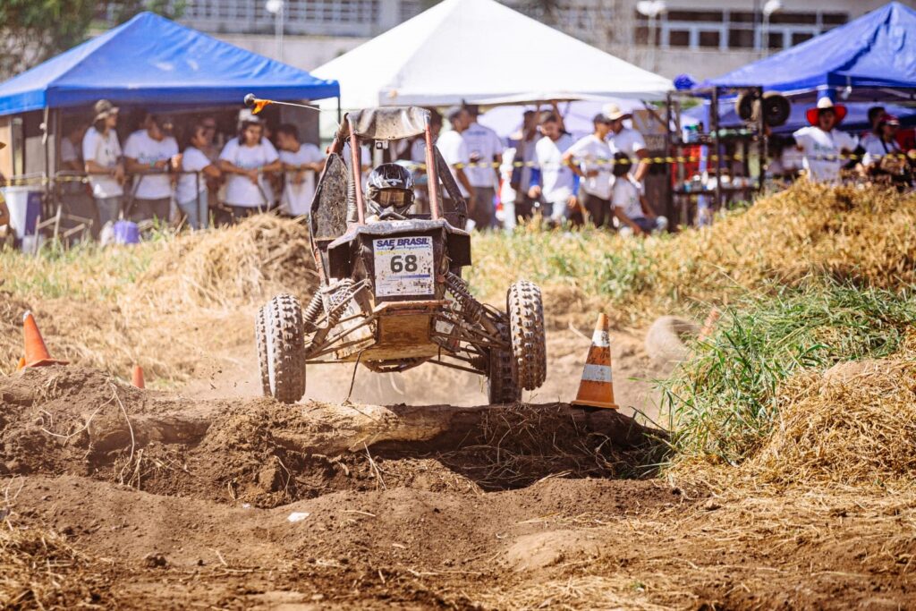 Um buggy de corrida, número 68, participa de um evento off-road. O veículo está em movimento, levantando terra e detritos enquanto atravessa um obstáculo de terra com um tronco de madeira. Um piloto com capacete protege o rosto do pó. Ao fundo, há espectadores sob tendas azuis e brancas, e muita vegetação seca e grama alta, indicando um ambiente de competição ao ar livre. Cones laranja marcam o percurso.