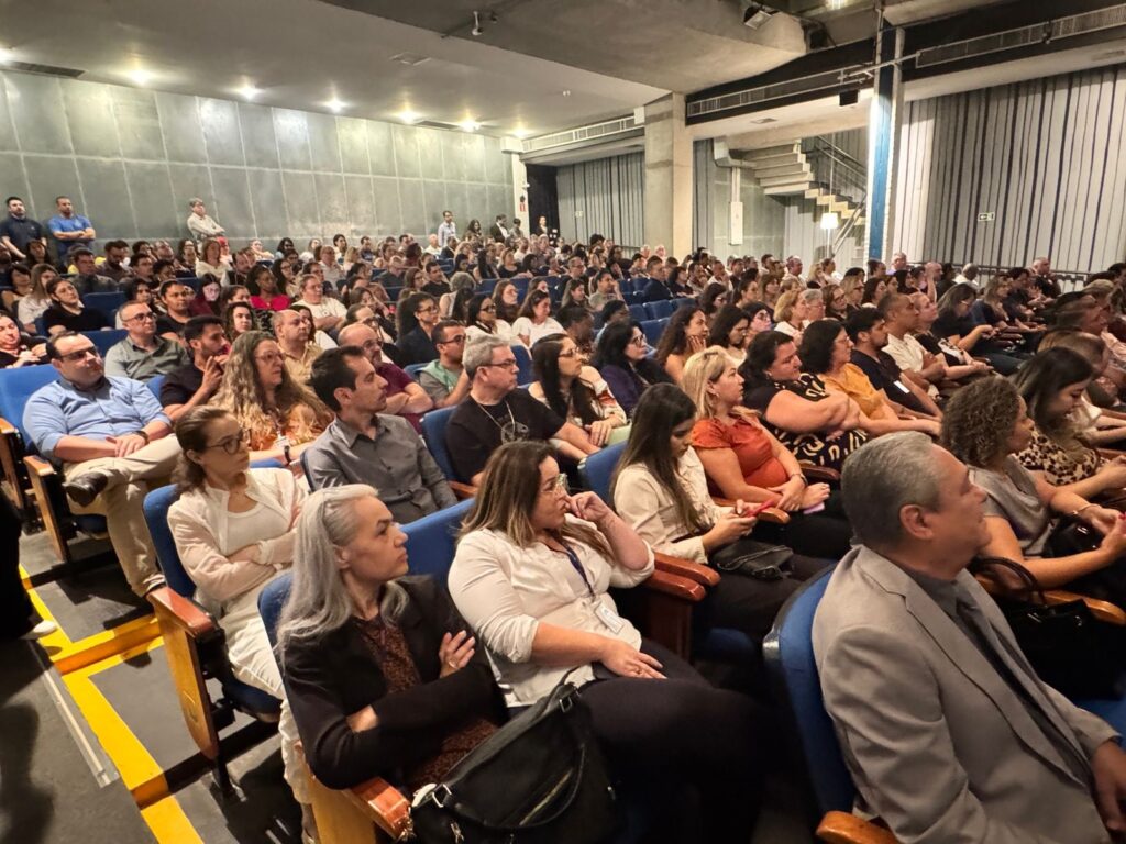 A imagem mostra uma plateia sentada em cadeiras azuis, voltada para a frente, como se estivesse assistindo a uma apresentação ou evento. As pessoas estão em diferentes posições, algumas olhando para a frente, outras conversando ou mexendo em seus celulares. O ambiente parece ser um auditório ou sala de conferências, com iluminação artificial que ilumina o teto e as paredes. Há uma escada visível ao fundo, sugerindo um espaço maior ou um edifício com múltiplos andares. A perspectiva da foto é de alguém sentado na plateia, olhando para os outros espectadores e para o palco ou área de apresentação.