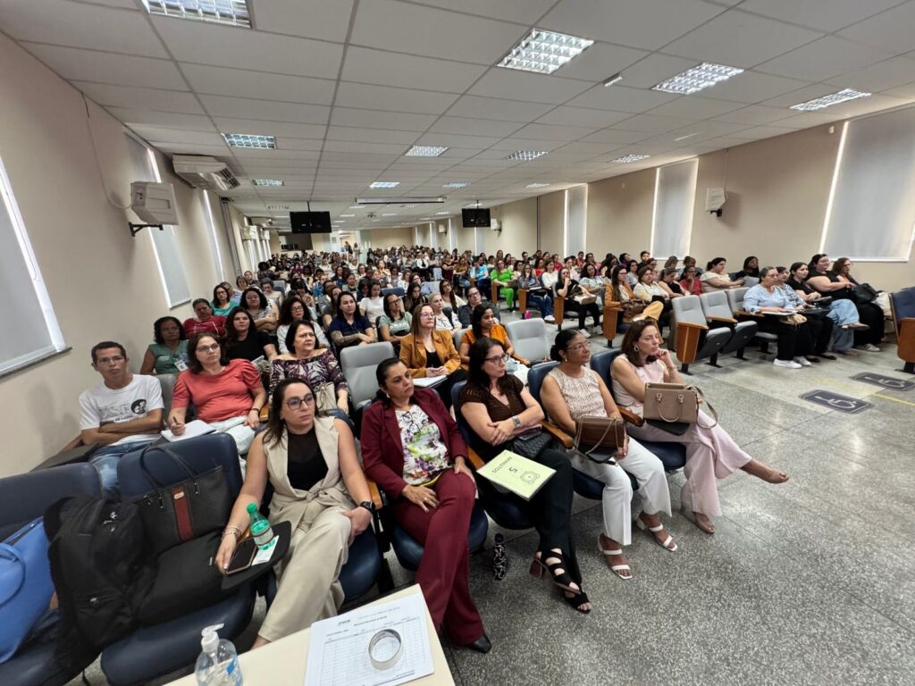 A imagem mostra uma sala de conferências cheia de pessoas sentadas em cadeiras, assistindo a uma apresentação. A maioria das pessoas são mulheres, e a sala parece ser um ambiente educacional ou profissional. As cadeiras estão dispostas em fileiras, voltadas para a frente da sala, onde provavelmente está o palestrante ou a tela de projeção. Há luminárias no teto e a iluminação é artificial. Algumas pessoas estão anotando, enquanto outras prestam atenção ao que está acontecendo. A atmosfera parece ser de aprendizado e engajamento.