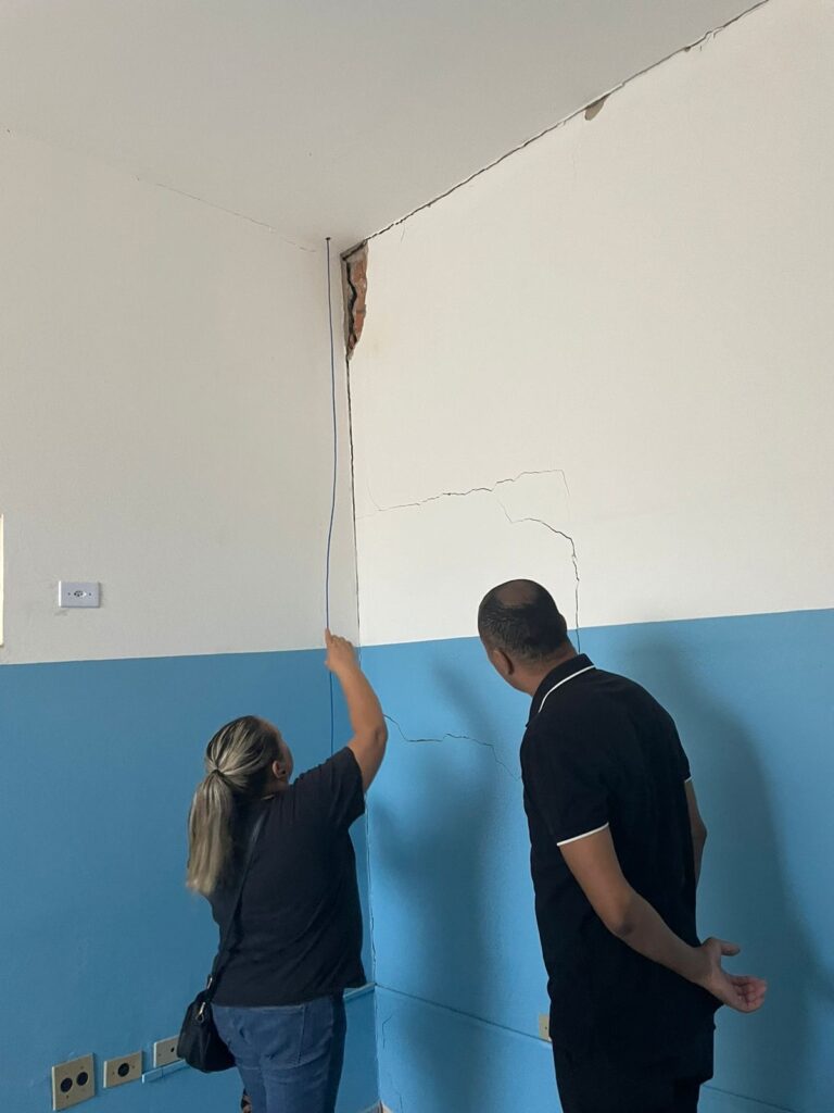 A imagem mostra duas pessoas em um ambiente interno, examinando uma parede com rachaduras visíveis. Uma mulher, vista de costas, aponta para uma rachadura vertical que se estende do teto até a metade da parede. A rachadura parece ter causado o desprendimento de parte do reboco, expondo a alvenaria de tijolos. Ao lado dela, um homem, também de costas, observa a mesma parede. A parede é dividida horizontalmente em duas seções: a superior é branca e a inferior é pintada de azul claro. Há também rachaduras horizontais e diagonais na seção branca da parede, abaixo da rachadura vertical principal. No canto inferior esquerdo, há tomadas elétricas na parede azul. A iluminação sugere que o ambiente é bem iluminado, possivelmente por luz natural entrando por uma janela ou por iluminação artificial.