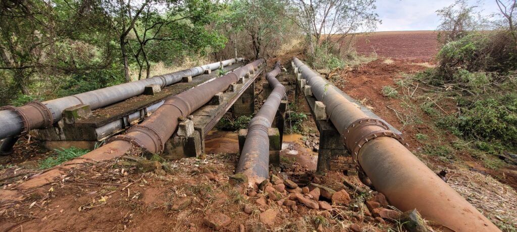 A imagem mostra uma série de tubulações grandes, algumas de metal enferrujado e outras de cor mais clara, dispostas sobre uma estrutura de concreto que atravessa um pequeno curso d'água. As tubulações parecem ser parte de um sistema de transporte de água ou outro fluido, e estão apoiadas em pilares de concreto que se elevam sobre o leito do riacho. Em alguns pontos, a água escorre pelas laterais das tubulações, sugerindo possíveis vazamentos ou gotejamentos. O entorno da estrutura é natural, com vegetação densa e árvores ao redor, criando uma atmosfera de um local mais isolado. O solo ao redor é de cor avermelhada, característico de terra argilosa, e há sinais de erosão e desmatamento em algumas áreas, especialmente no lado direito da imagem, onde uma encosta inclinada com terra exposta e galhos cortados é visível. A combinação de infraestrutura industrial com o ambiente natural sugere uma paisagem rural ou de transição entre o desenvolvimento humano e a natureza.