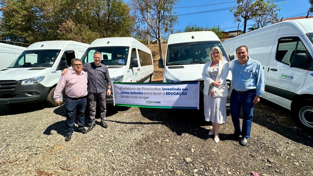 A imagem mostra quatro vans brancas estacionadas em um terreno com cascalho, sob luz solar intensa. Três das vans estão totalmente visíveis, enquanto uma quarta está parcialmente visível à esquerda.No centro da imagem, um banner branco com letras azuis e verdes está estendido na frente das vans. O banner diz: "Prefeitura de Piracicaba: investindo em novos veículos para levar a EDUCAÇÃO ainda mais longe!". Abaixo deste texto, há um logotipo menor com as palavras "PIRACICABA" e "CUIDANDO DO QUE PRECISA SER FEITO".Quatro pessoas estão em pé em frente às vans e ao banner. À esquerda, dois homens estão juntos, com um deles com a mão no ombro do outro. O homem da esquerda usa uma camisa rosa clara e jeans escuros, enquanto o homem da direita usa uma camisa cinza escura e calças cinzas escuras.À direita, um homem e uma mulher estão em pé. O homem usa uma camisa azul clara de botões e jeans escuros. A mulher usa um vestido cinza claro com babados e um blazer branco.