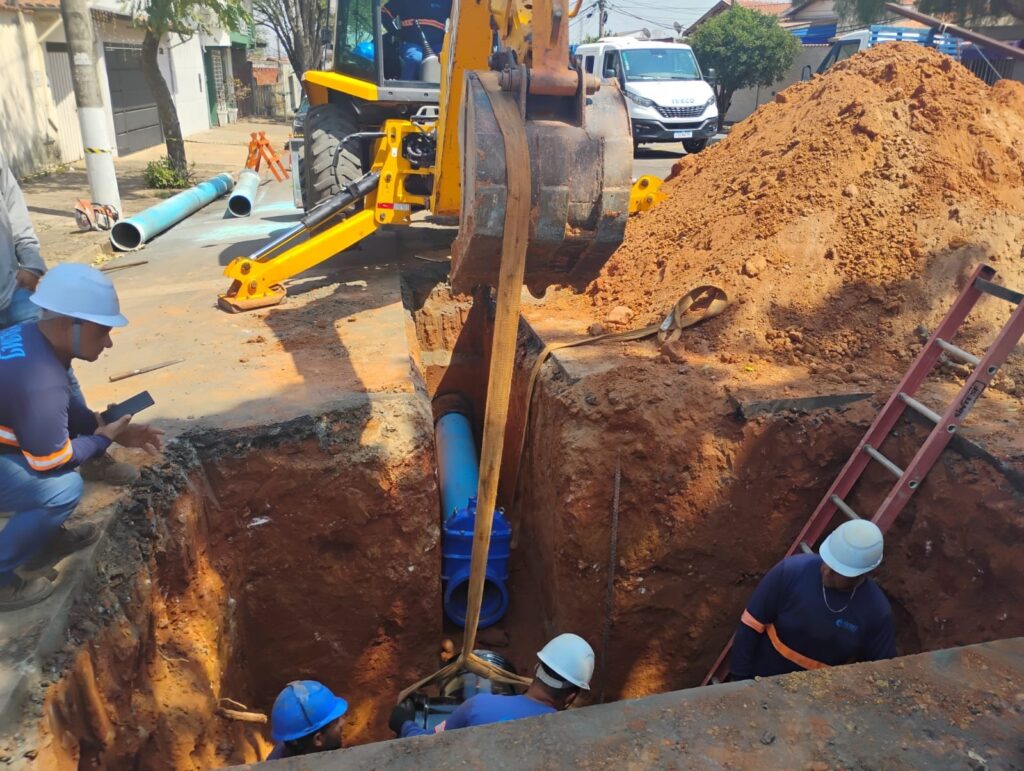 A imagem mostra uma equipe de trabalhadores em uma vala, instalando uma tubulação de água. Três homens, todos usando capacetes e uniformes de trabalho, estão envolvidos no processo. Um homem, com um capacete branco, está de pé e usando uma fita métrica para medir uma peça de tubulação preta que está sendo suspensa por uma linga amarela. Outro homem, com um capacete azul, está agachado e ajudando a manusear a tubulação preta. Um terceiro homem, também com um capacete branco, está agachado e parece estar supervisionando o trabalho ou auxiliando na medição. Acima deles, uma tubulação azul maior, com uma válvula azul proeminente, está instalada na vala. A vala em si é profunda, com paredes de terra marrom e laranja, e há sinais de trabalho de construção, como pedras e detritos. Uma escada vermelha está encostada em uma das paredes da vala, sugerindo o acesso ao local. O sol parece estar brilhando, criando sombras na vala.
