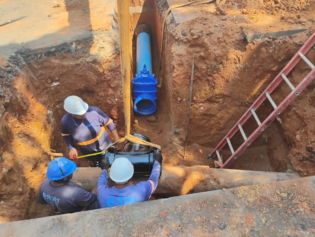A imagem mostra uma equipe de trabalhadores em uma obra de saneamento básico, possivelmente substituindo ou reparando uma tubulação de água. * No centro da imagem, um escavadeira amarela está posicionada sobre uma vala profunda, com sua concha levantando uma nova tubulação azul. * Abaixo da concha, a vala expõe a tubulação antiga e uma válvula azul de grande porte, indicando a importância do trabalho. * Vários trabalhadores, vestidos com uniformes de segurança e capacetes, estão na vala e ao redor dela, supervisionando e auxiliando no processo. Um deles está ajoelhado e olhando para um celular, possivelmente verificando plantas ou instruções. * Uma escada vermelha está apoiada na lateral da vala, permitindo o acesso dos trabalhadores. * Ao fundo, observa-se uma rua com algumas casas e um veículo branco da marca Iveco, sugerindo que a obra está ocorrendo em uma área urbana. * Há montes de terra removida ao redor da vala, evidenciando a extensão da escavação.