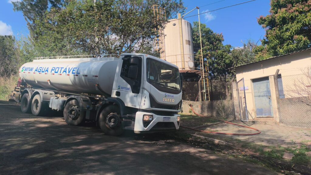 A imagem mostra um caminhão de água branco estacionado em uma estrada de terra. O caminhão tem "SEMAE ÁGUA POTÁVEL" escrito na lateral, indicando que é para distribuição de água potável pelo SEMAE. O modelo do caminhão é IVECO, como pode ser visto na parte da frente. Ao fundo, há um tanque de água cilíndrico e alto, provavelmente parte de um sistema de abastecimento de água. Uma escada de metal está anexada ao tanque para acesso. Perto do tanque, há um pequeno edifício de cor clara com uma porta azul e uma cerca de arame em volta da área. Uma mangueira vermelha é visível, indo da proximidade do caminhão em direção ao edifício. A cena parece estar em um ambiente rural ou semi-rural, com árvores e vegetação ao redor da infraestrutura de abastecimento de água. O céu está azul com algumas nuvens, sugerindo um dia claro.