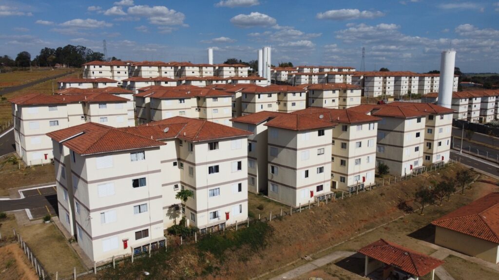 A imagem mostra um grande conjunto habitacional com vários prédios de apartamentos idênticos. Os prédios são de cor creme com detalhes em marrom e telhados de cerâmica vermelha. Eles têm várias janelas com persianas brancas e portas de entrada vermelhas. Os prédios estão dispostos em fileiras em uma paisagem levemente inclinada, com algumas árvores e vegetação espalhadas. Ao fundo, há duas torres brancas altas, provavelmente torres de água ou de comunicação. O céu está azul com algumas nuvens brancas fofas, indicando um dia claro e ensolarado. A cena geral sugere um empreendimento residencial moderno e bem cuidado.