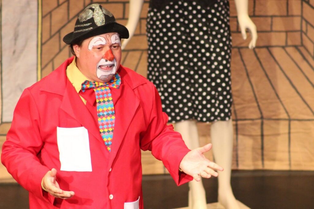 A imagem mostra um palhaço em close-up, vestindo uma roupa vibrante e maquiagem característica. Ele usa um chapéu preto com detalhes brilhantes, uma jaqueta vermelha aberta sobre uma camisa amarela e uma gravata borboleta colorida com um padrão quadriculado. Seu rosto está pintado com branco, com círculos vermelhos nas bochechas e um nariz vermelho. As mãos do palhaço estão estendidas para a frente, com as palmas para cima, em um gesto convidativo ou de apresentação. Ao fundo, desfocado, aparece a parte inferior do corpo de uma figura feminina vestindo uma saia preta com bolinhas brancas e pernas de manequim, sugerindo um cenário de palco ou apresentação. O fundo atrás do palhaço e da figura feminina parece ser uma representação estilizada de uma parede de tijolos e um piso de madeira.
