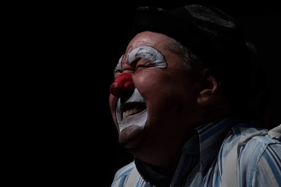 A imagem é um close-up de um palhaço com o rosto pintado, virado para a direita numa pose dramática. O palhaço tem os olhos fechados, com a testa franzida e a boca aberta num grito ou risada expressiva, revelando os dentes. A sua maquilhagem inclui uma base branca na testa e em volta dos olhos, um nariz vermelho brilhante e uma área prateada em volta da boca e do queixo. O palhaço usa um chapéu preto em cima da cabeça e uma camisa listrada de azul e branco. A iluminação é dramática, com uma luz forte a incidir sobre o lado direito do rosto do palhaço, criando sombras profundas no lado esquerdo e no fundo, que é completamente preto. Esta iluminação realça as texturas da pele, a maquilhagem e a expressão emocional do palhaço.