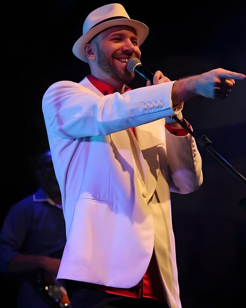 A imagem exibe um cantor em um palco, iluminado por luzes coloridas que criam um ambiente vibrante. Ele veste um terno branco impecável, um chapéu estilo fedora e uma camisa vermelha, compondo um visual elegante e chamativo. O cantor está com um sorriso contagiante, segurando um microfone e apontando com a mão direita, transmitindo energia e interação com o público. Ao fundo, desfocado, um músico toca um instrumento, sugerindo a presença de uma banda que acompanha a apresentação. A iluminação destaca o cantor, criando um jogo de luzes e sombras que adiciona profundidade à cena.