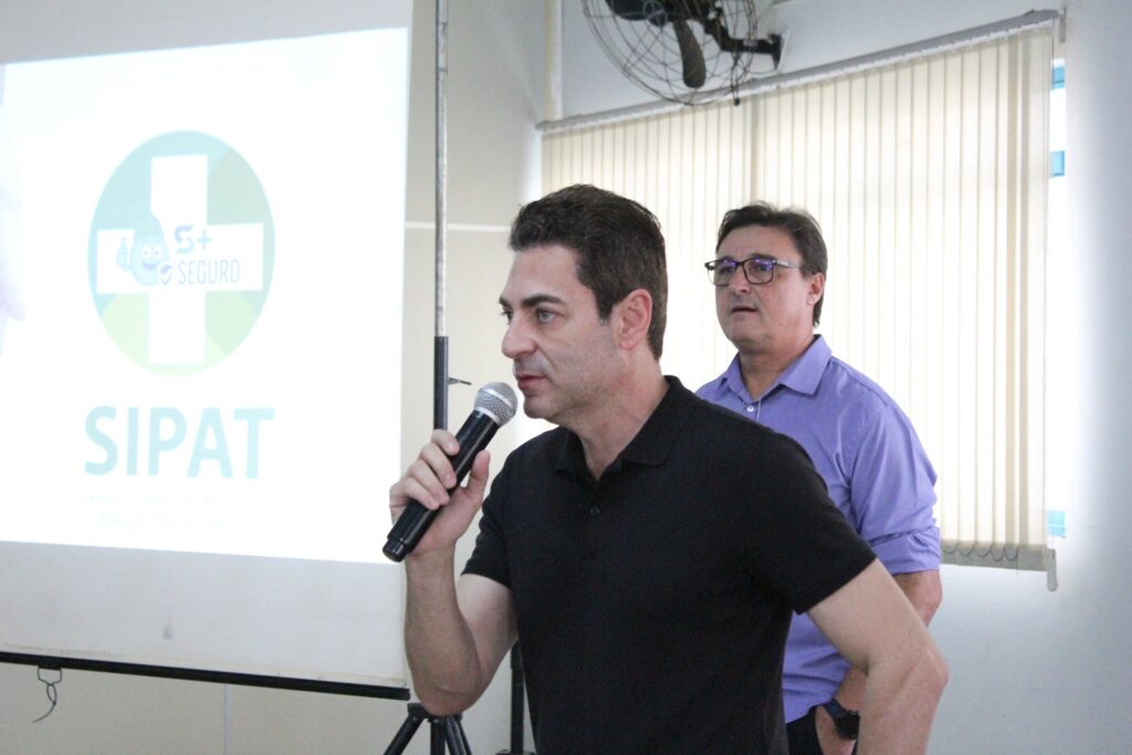 A imagem mostra dois homens em uma sala, possivelmente durante uma apresentação ou evento. O homem em primeiro plano, à esquerda, está de perfil, segurando um microfone e falando. Ele veste uma camisa preta de gola polo. Atrás dele, à direita, está outro homem, olhando para a frente, vestindo uma camisa roxa e óculos. Ao fundo, uma tela de projeção exibe um logotipo com um símbolo de cruz verde e um personagem sorridente com capacete, além do texto "SIPAT". As janelas ao fundo estão cobertas por persianas verticais.