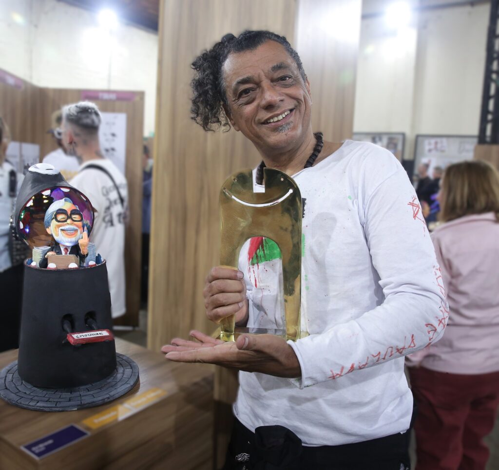 Rui Miranda sorri segurando troféu dourado, ao lado de sua escultura colorida exposta.