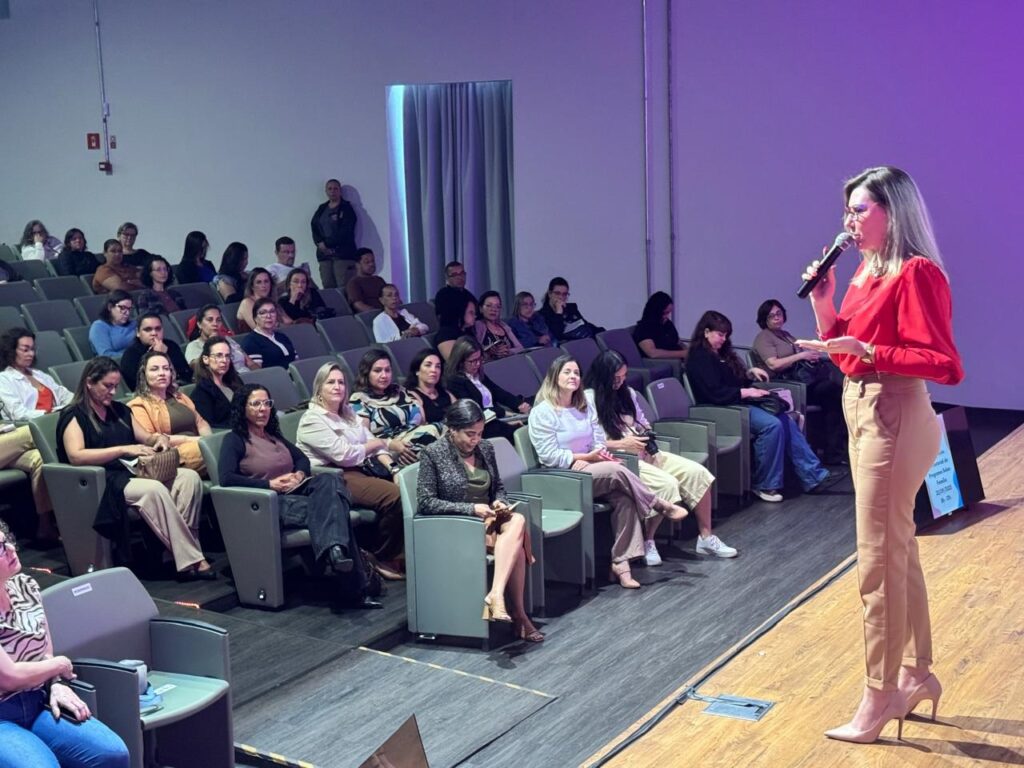 A imagem mostra uma sala de conferências com uma plateia sentada em cadeiras dispostas em arquibancadas, voltada para um palco. Uma mulher está em pé no palco, segurando um microfone e falando para a plateia. Ela está vestida com uma blusa vermelha e calças bege. A plateia parece composta por adultos, com predominância de mulheres. O ambiente é iluminado e parece ser uma apresentação ou palestra.