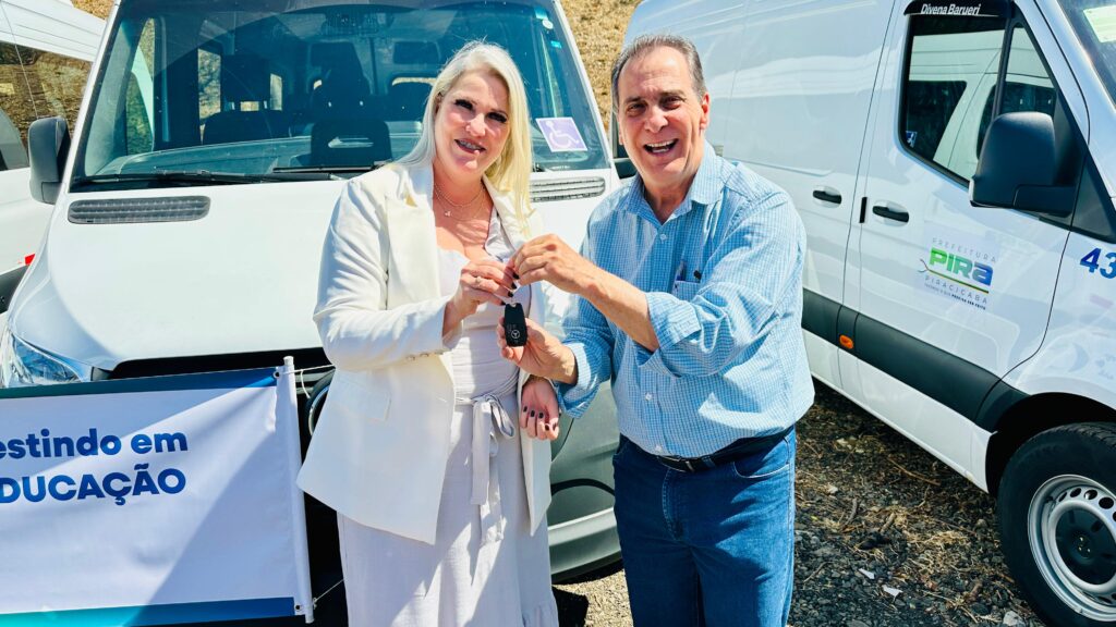 Na imagem, uma mulher e um homem estão em frente a uma van branca. A mulher, com cabelos loiros e usando um blazer branco, segura as chaves de um carro na mão direita, enquanto o homem, vestindo uma camisa xadrez azul e jeans, estende a mão para pegá-las. Ambos sorriem para a câmera.