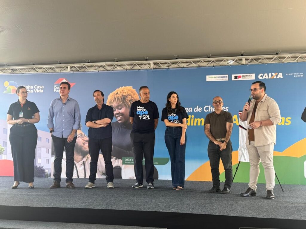 A imagem mostra um grupo de sete pessoas em pé em um palco, com um banner ao fundo. As pessoas parecem estar participando de um evento oficial, possivelmente relacionado a habitação ou desenvolvimento social, dado o texto no banner. Do lado esquerdo, há uma mulher com um colete preto e calças pretas, parecendo ser a primeira a falar ou apresentar. Ao lado dela, um homem de camisa azul clara e calças escuras, com as mãos nos bolsos. Em seguida, um homem de camisa escura e calças escuras, com os braços cruzados. O quarto indivíduo é um homem com uma camiseta preta com a inscrição "Meu novo ape SPL". No lado direito do palco, há uma mulher com uma camiseta preta com a mesma inscrição "Meu novo ape SPL", usando calças jeans. Ao lado dela, um homem de camisa marrom e calças escuras, com as mãos atrás das costas. O último homem à direita está de camisa bege clara e calças bege, segurando um microfone e falando, aparentemente o orador principal. O banner ao fundo exibe logotipos de instituições como "CAIXA", "SÃO PAULO" e "MINISTÉRIO DAS CIDADES", além de texto em português que inclui "Minha Casa Minha Vida" e "Piracicaba". Há também uma imagem estilizada de um prédio e um gráfico colorido no canto inferior direito. A iluminação sugere que o evento está ocorrendo durante o dia, ao ar livre ou em um local com boa iluminação natural.