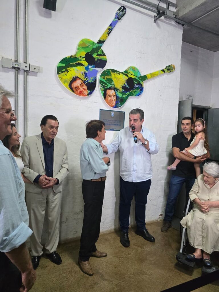 Na imagem aparece um senhor de bigode segurando um microfone em sua mão esquerda. Ela olha e está com o braço estendido no ombro de um senhor de camisa clara e calça escura, que olha atentamente para ele. Ao lado deles aparecem quatro pessoas em pé, sendo um rapaz segurando uma criança no colo. E ao lado está uma senhora de cabelos brancos, que olha cabisbaixo. Na parede estão o desenho de dois violões, estampado com o rosto de dois homens.