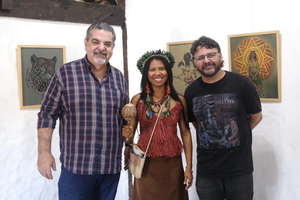 Na imagem, três pessoas posam juntas em frente a quadros. À esquerda, um homem de meia-idade, com cabelos grisalhos e barba, veste uma camisa xadrez azul e branca e jeans. Ele sorri para a câmera. No centro, uma mulher com trajes tradicionais indígenas, incluindo um cocar de penas verdes e vermelhas, colares e uma blusa de franjas, segura um chocalho na mão direita e uma bolsa de palha a tiracolo. Ela também sorri para a câmera. À direita, um homem com óculos e barba veste uma camiseta preta com a estampa da banda "The Smiths" e calça jeans escura. Ele está levemente inclinado para a frente, com as mãos atrás das costas. Ao fundo, observam-se três quadros com ilustrações. O quadro à esquerda exibe o retrato de uma onça pintada. O quadro do meio apresenta figuras humanas em um estilo mais abstrato. O quadro à direita mostra uma representação estilizada de uma figura feminina sob um sol radiante. As paredes são brancas e o teto é de madeira escura.