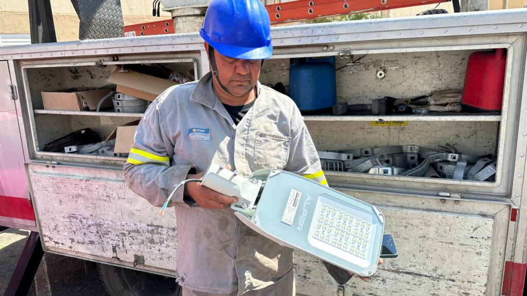 Um trabalhador com capacete azul e uniforme de trabalho cinza está segurando uma luminária de LED branca em suas mãos. A luminária tem uma longa fileira de LEDs brancos brilhantes. Atrás dele, há um caminhão com uma caixa de ferramentas aberta, revelando vários compartimentos cheios de equipamentos e suprimentos. Vê-se uma escada vermelha no topo da caixa de ferramentas.