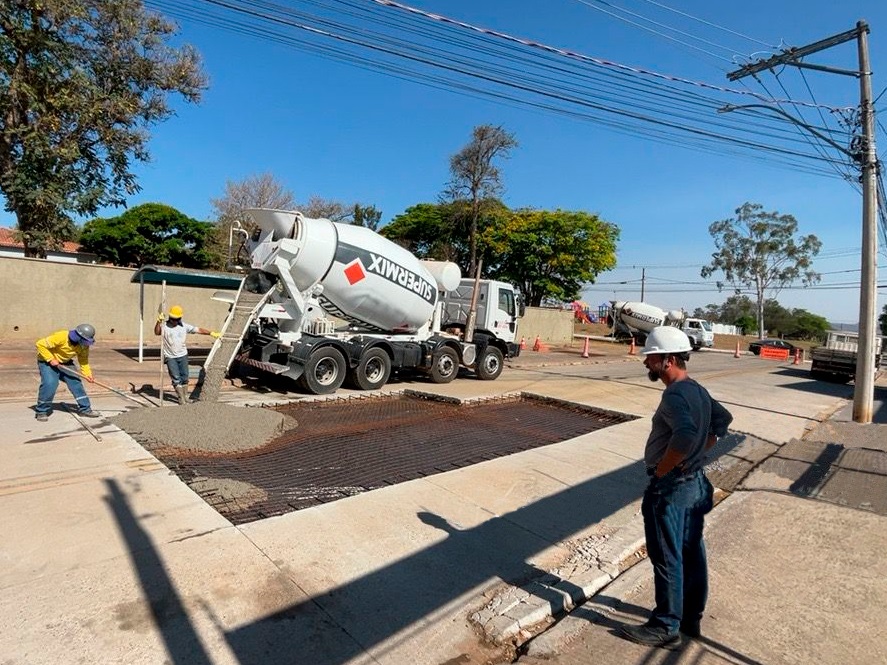 A imagem mostra uma cena de construção em uma rua ensolarada, com um caminhão betoneira da "SUPERMIX" descarregando concreto em uma área preparada com malha de aço. Dois trabalhadores estão envolvidos no processo: um, vestindo um capacete amarelo e uma jaqueta amarela, manobra o concreto com uma ferramenta, enquanto outro, com capacete branco e camiseta branca, parece supervisionar. Ao fundo, outro caminhão betoneira está estacionado, e cones de trânsito indicam uma área de trabalho. Um terceiro trabalhador, com capacete branco e camiseta azul escura, observa a cena de pé na calçada. A imagem captura um momento de atividade em uma obra de pavimentação, com o céu azul claro e árvores ao fundo, sugerindo um dia de bom tempo.