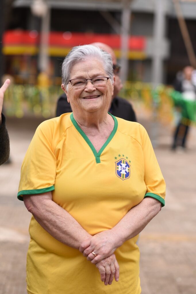 Na imagem aparece uma senhora de óculos e cabelos brancos, sorrindo. Ela de braços cruzados, veste uma camiseta amarela e no lado esquerdo está escrito CBF.