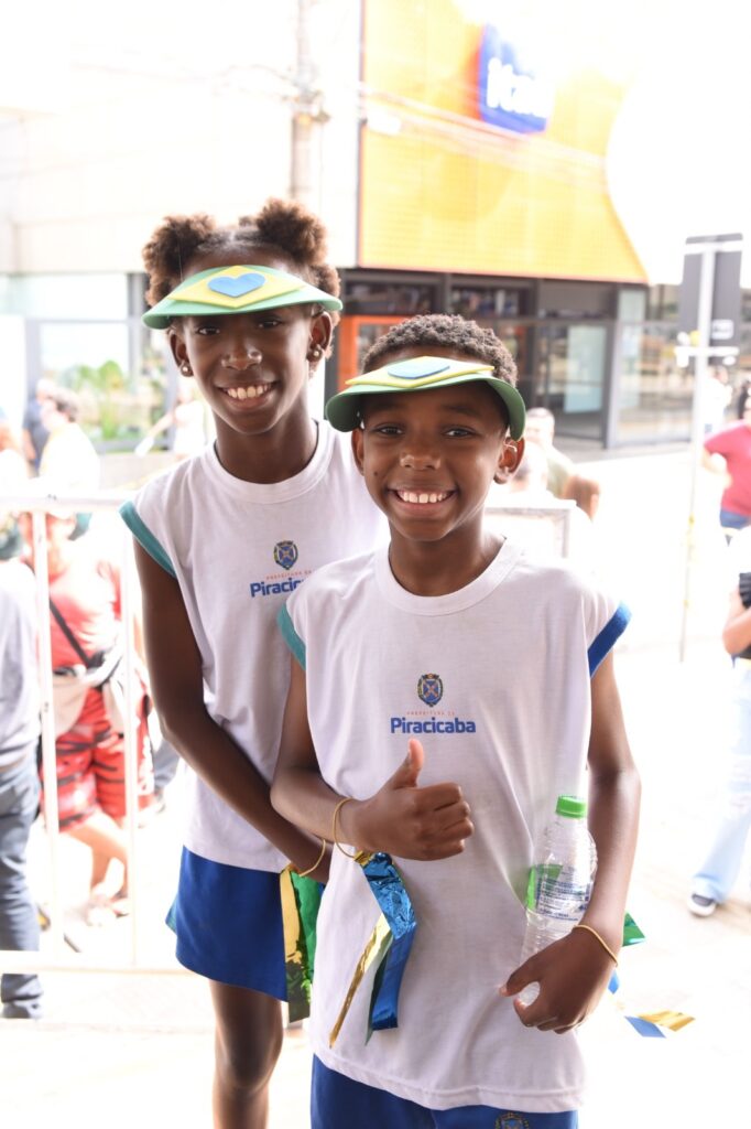 Na imagem aparece uma menina e um menino sorrindo. Eles vestem o uniforme composto de camiseta branca escrito Piracicaba ao meio e acima um brasão e shorts azul marinho. Ambos vestem uma proteção na cabeça, nas cores amarelo, azul e verde. O menino traz em sua mão esquerda uma garrafinha de água.