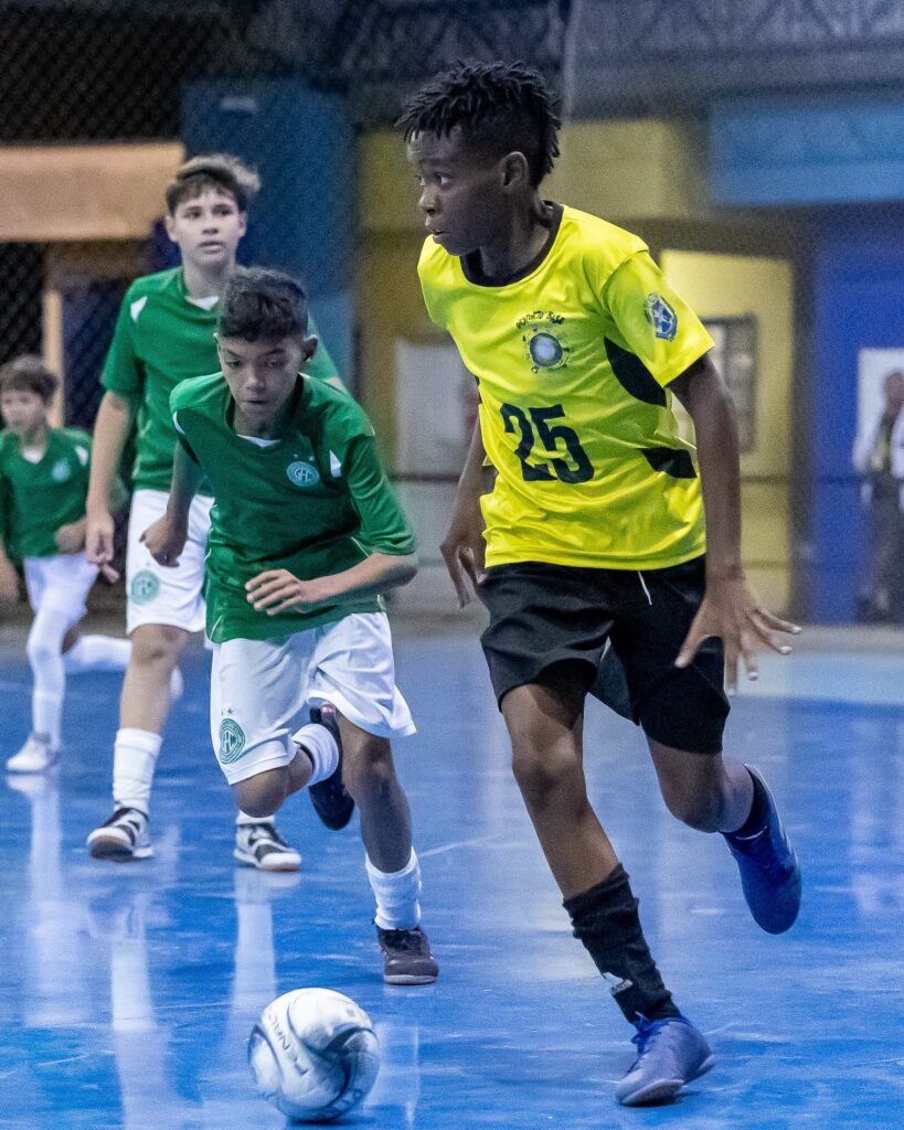 No primeiro plano, à direita, um menino com pele escura e cabelo curto e crespo corre em direção à câmera, vestindo uma camisa amarela com o número 25 estampado e calções pretos. Ele está com a perna direita esticada para a frente, e a esquerda ligeiramente dobrada, em plena ação de corrida. Seu olhar está focado para a frente, com uma expressão de concentração. À sua esquerda, um pouco mais atrás, um menino com pele clara e cabelo escuro e curto também corre, vestindo uma camisa verde com detalhes brancos e calções brancos. Ele está com a perna esquerda à frente e a direita um pouco atrás, também em movimento. Seu olhar está direcionado para o chão, possivelmente acompanhando a bola. Ao fundo, mais atrás e à esquerda, outro menino, com pele clara e cabelo castanho, veste uma camisa verde semelhante à do menino à sua esquerda. Ele está em segundo plano, também em movimento. No chão, em primeiro plano e ligeiramente à esquerda do centro, uma bola de futebol de salão branca com detalhes pretos está parada. O piso da quadra é azul, refletindo a luz e as figuras dos jogadores. O fundo da imagem é escuro, com algumas luzes e contornos desfocados de pessoas e estruturas, sugerindo um ambiente interno, como um ginásio.