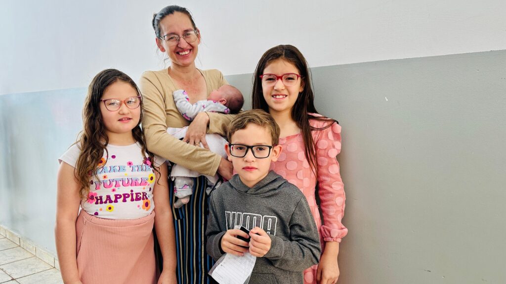 A imagem mostra uma família posando para uma foto em um corredor. Há uma mulher adulta no centro, segurando um bebê recém-nascido. Ela está usando óculos e um suéter bege. À sua esquerda está uma menina com cabelos longos e escuros, usando óculos e uma camiseta branca com a frase "MAKE THE FUTURE HAPPIER" estampada, combinada com uma saia rosa. À direita da mulher, está uma menina mais velha com cabelos longos escuros, usando óculos vermelhos e um vestido rosa com bolinhas. Na frente, há um menino usando óculos pretos e um moletom cinza. Ele está segurando um pequeno objeto preto e um pedaço de papel. O fundo é uma parede dividida em duas cores: branco na parte superior e cinza na parte inferior.