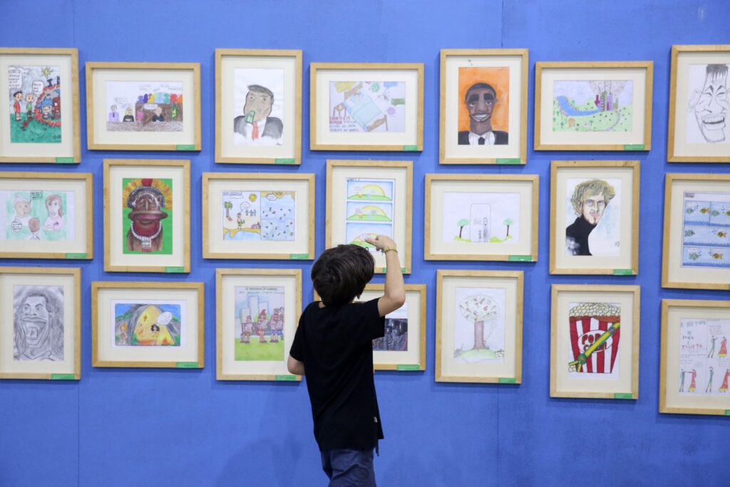 Menino observa desenhos infantis emoldurados expostos em parede azul no Salãozinho de Humor.