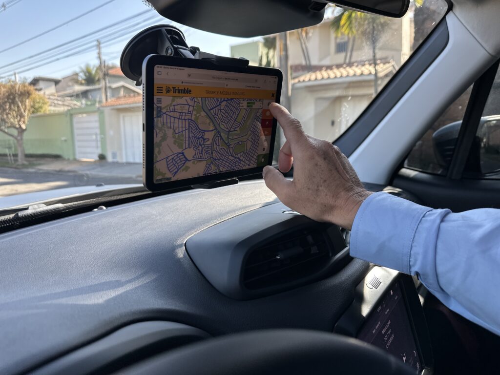 Na imagem, vemos um close-up do interior de um carro, com foco em um tablet posicionado no para-brisa. O tablet exibe um mapa detalhado com marcações e a logo "Trimble" no canto superior esquerdo, juntamente com o título "TRIMBLE MOBILE IMAGING". Uma mão com uma camisa azul clara de mangas compridas está tocando a tela do tablet, especificamente um ícone no lado direito que se assemelha a uma engrenagem. A luz do sol ilumina o painel do carro e o exterior, indicando que é dia. Ao fundo, vislumbramos casas com telhados e árvores, sugerindo um ambiente residencial. Parte do painel do carro, incluindo uma saída de ar, e o volante também são visíveis, emoldurando a cena. O tablet está preso ao para-brisa por um suporte com ventosa. Na parte inferior direita da tela do tablet, há informações de áudio com o título "Música pop" e detalhes de estação e artista.