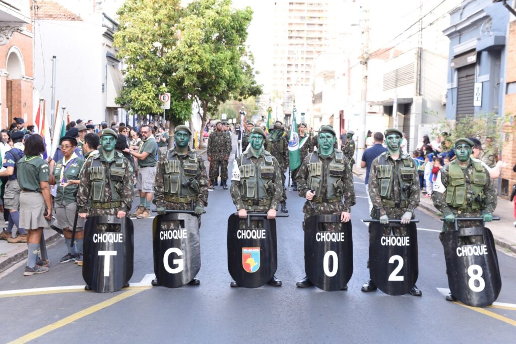 Na imagem aparecem à frente do desfile, seis policiais camuflados, vestindo boinas e uniforme verde mata. A frente seguram um escudo preto em que está escrito Choque e em baixo TG 028