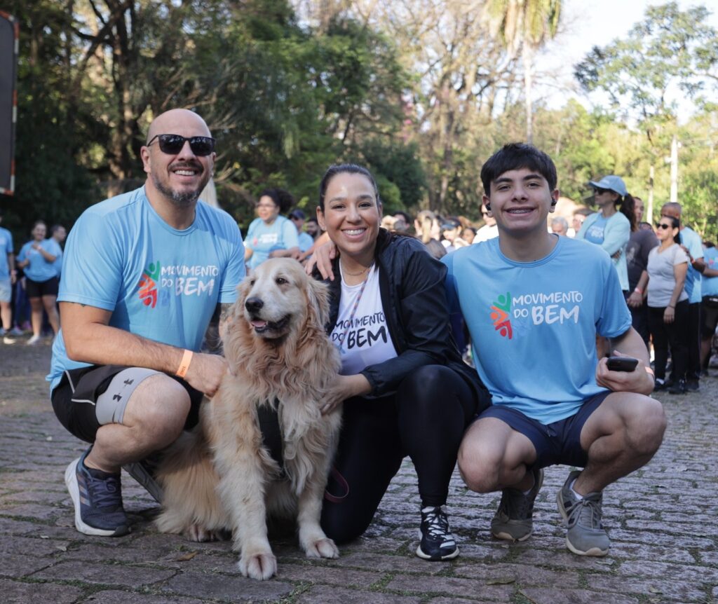 A imagem mostra quatro pessoas sorrindo, em frente a um banner com a inscrição "MOVIMENTO DO BEM". Duas mulheres estão à esquerda, vestindo camisetas brancas com o logo do movimento. A mulher mais à esquerda usa óculos de sol escuros e a outra usa um colar. Uma mulher no centro está vestindo um top preto e leggings com um padrão degradê, um boné e óculos escuros. Um homem está à direita, vestindo uma camiseta azul clara com o logo do movimento, óculos escuros e mangas compridas pretas. Ele está segurando o selim de uma bicicleta preta que está posicionada no centro da imagem. O fundo sugere um evento ao ar livre, com árvores e outras pessoas ao fundo.