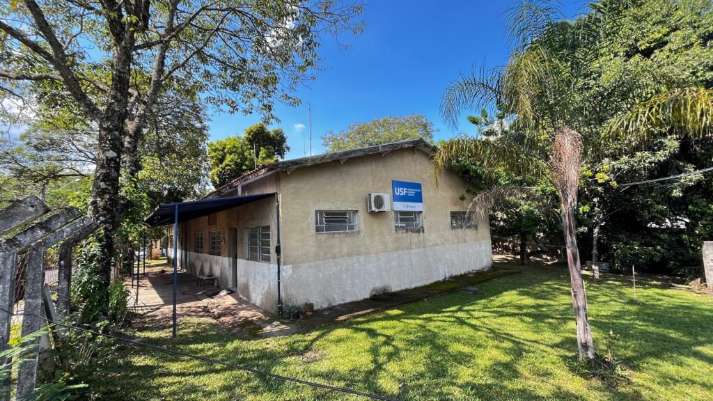 A imagem mostra um prédio com um telhado de telhas marrons e paredes de cor bege claro. Há uma placa azul com o logo "USF" e texto em branco no lado direito da fachada. Um aparelho de ar condicionado está instalado acima de uma das janelas retangulares. A frente do prédio é protegida por um toldo azul. O prédio está cercado por vegetação exuberante, com árvores altas e grama verde bem cuidada. Uma cerca de concreto com arame farpado está visível no lado esquerdo da imagem. O céu está azul claro com algumas nuvens brancas dispersas. A iluminação sugere que é um dia ensolarado.