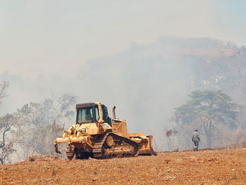 A imagem mostra um trator amarelo em uma encosta com vegetação rasteira e algumas árvores secas. O trator está posicionado à esquerda do quadro, voltado para a direita. Um homem vestindo uniforme caminha para longe do trator, em direção à direita do quadro. O fundo está obscurecido por uma densa névoa ou fumaça, que adiciona uma atmosfera etérea à cena. A fumaça parece emanar de uma área mais distante, possivelmente de uma queimada ou incêndio florestal, obscurecendo as colinas e a vegetação mais afastadas. O céu está nublado e acinzentado, contribuindo para a atmosfera geral de neblina. A iluminação sugere um dia nublado ou com pouca luz solar.