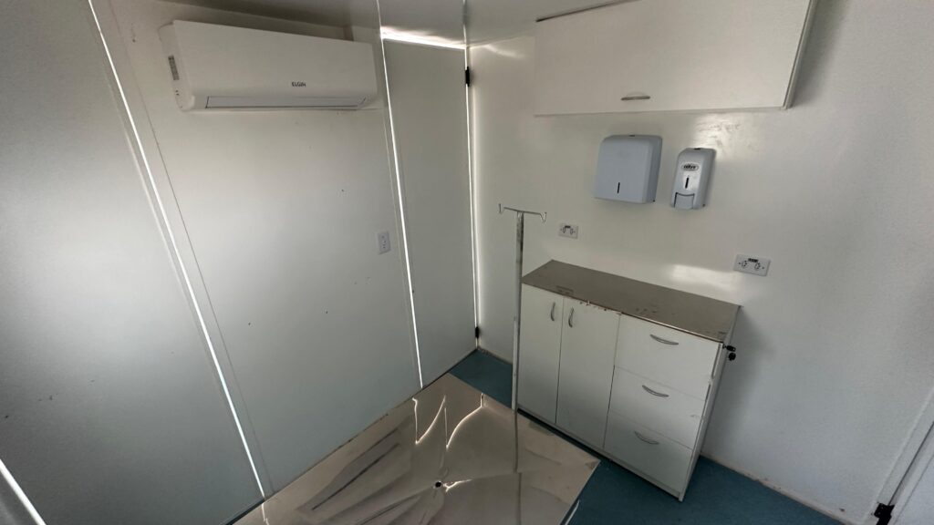 A imagem mostra: * Uma sala com paredes, piso e teto brancos. * Ao fundo, uma porta branca entreaberta, permitindo a entrada de luz. * À esquerda, um ar condicionado Elgin instalado na parede. * No centro da sala, uma mesa de exame de metal com um suporte para soro ao lado. * À direita, um armário branco com tampo de metal e gavetas, e acima dele, dois dispensadores na parede: um para papel toalha e outro para álcool em gel. * Há também tomadas elétricas visíveis nas paredes.