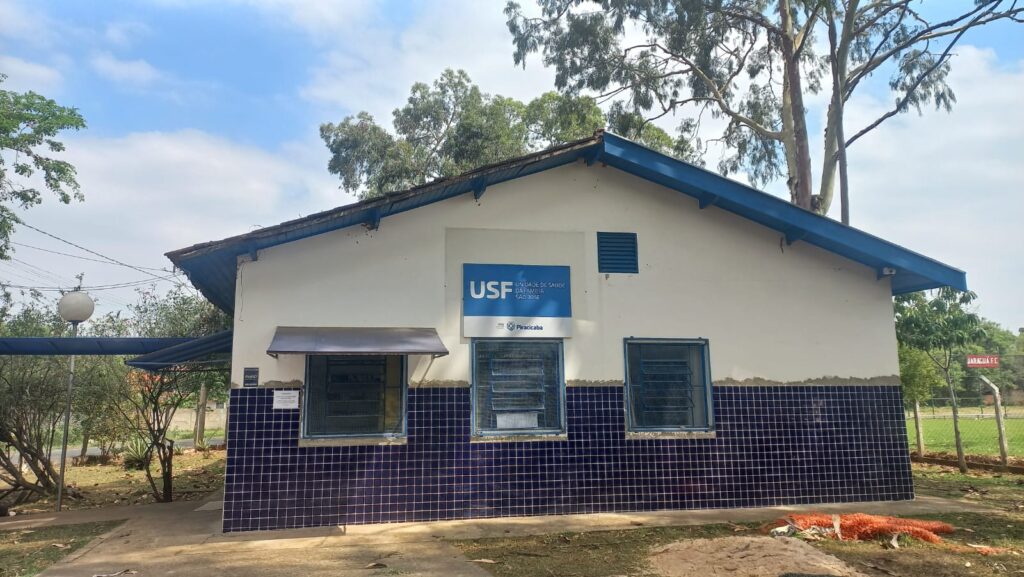 A imagem mostra a fachada de uma edificação, provavelmente uma unidade de saúde, com um céu azul e nuvens ao fundo. A construção possui um telhado azul inclinado, paredes brancas e uma base revestida com azulejos azuis quadriculados. Há três janelas visíveis na fachada, sendo que duas delas estão protegidas com grades. Uma placa com o logo "USF" e o nome "UNIDADE DE SAUDE DA FAMILIA SAO JOSE" está fixada na parede, acima de uma janela central. Abaixo desta placa, há outra com o nome "Piracicaba". Uma pequena marquise de metal cobre a janela mais à esquerda. Ao redor da edificação, observa-se vegetação, incluindo árvores com folhas verdes e galhos secos, e alguns materiais de construção no chão, como uma rede laranja. A presença de uma cerca e um campo gramado ao fundo sugere que a edificação está localizada em uma área mais aberta, possivelmente um bairro ou um espaço comunitário.
