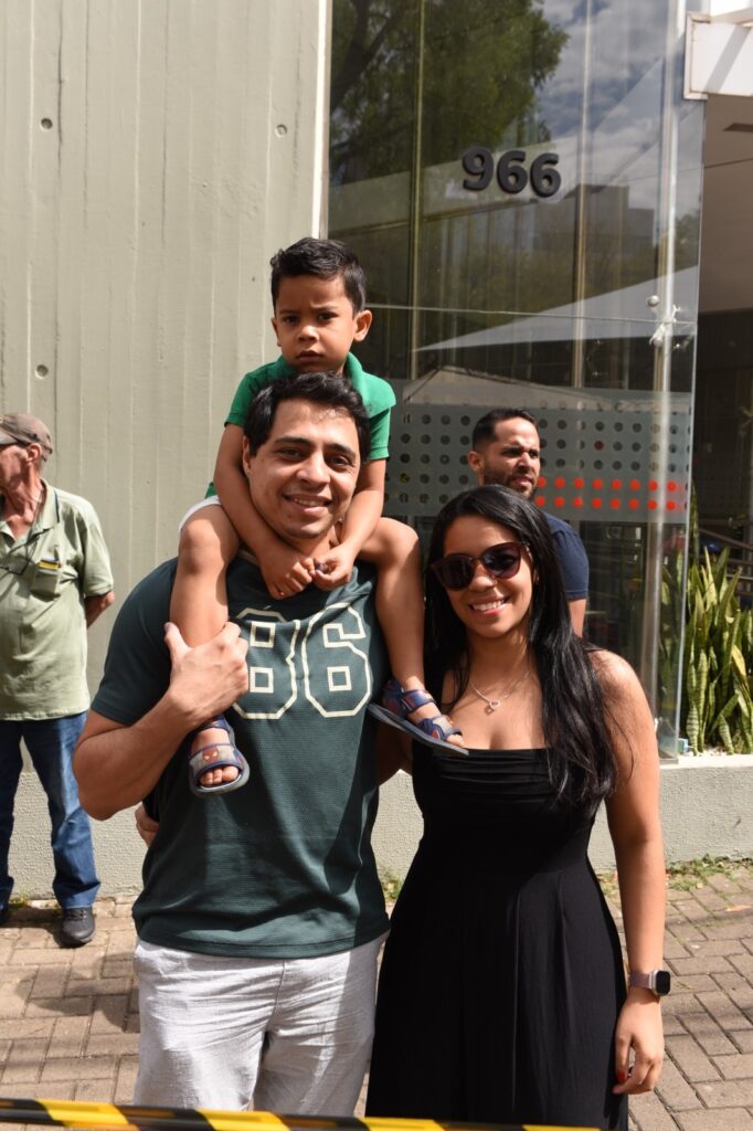 Na imagem aparece um casal. O homem veste camiseta verde e bermuda clara, carregando um menino de camiseta verde, short branca e chinelinho azul comdetqalhes em vermelho. Ao seu lado aparece uma moça de cabelos pretos e óculos escuro, sorridente, vestindo um vestido preto. Ao fundo aparece um edifício com o número 966.