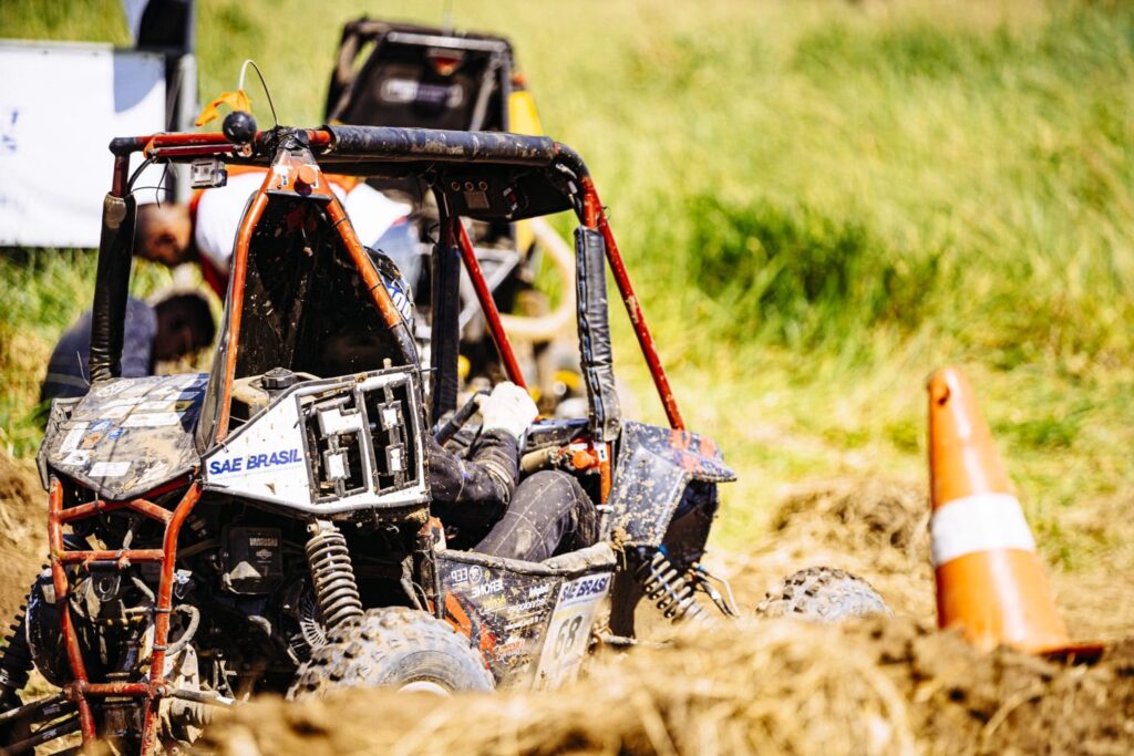 A imagem mostra um veículo off-road sujo de lama, em um campo de grama verde e solo seco. * O veículo, possivelmente um buggy de competição, tem uma estrutura tubular vermelha e preta e parece estar atolado na lama. * Um pneu com sulcos profundos é visível, assim como a suspensão robusta. * Há adesivos no veículo, incluindo um que diz "SAE BRASIL" e o número "68" em destaque. * Uma pessoa com luvas e traje de corrida está sentada no banco do motorista, com as mãos no volante. * Ao fundo, outros veículos semelhantes e algumas pessoas são visíveis, sugerindo um evento ou competição de off-road. * Um cone de trânsito laranja com uma faixa branca está posicionado na lateral direita da imagem, indicando um obstáculo ou limite. * A iluminação sugere que a foto foi tirada em um dia ensolarado.