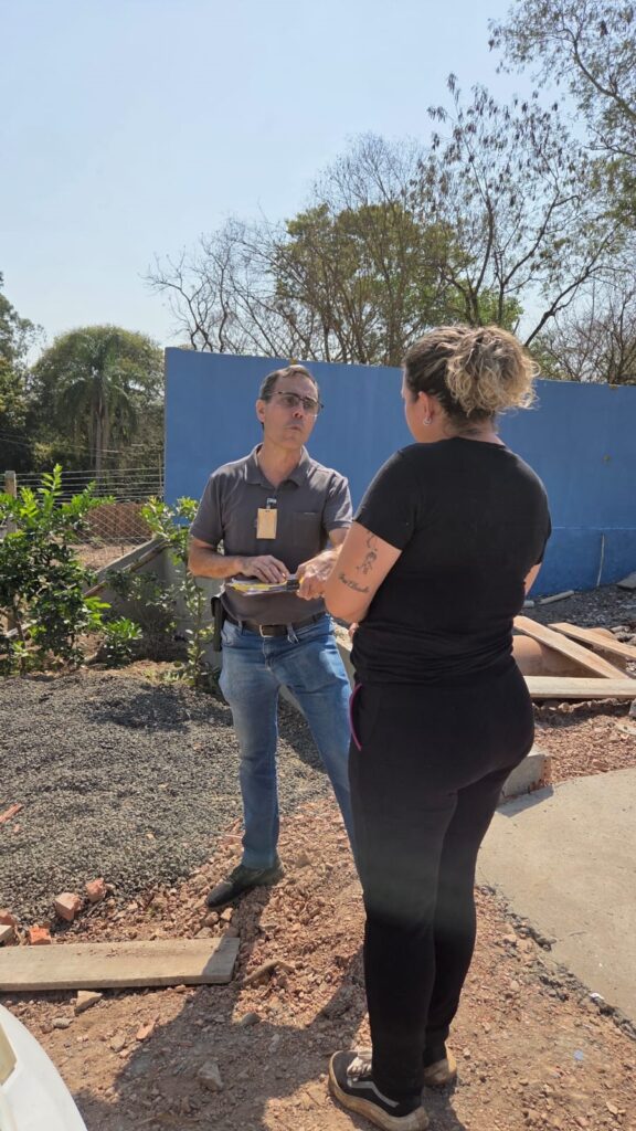 A imagem mostra duas pessoas conversando ao ar livre, em um dia ensolarado. Um homem, vestindo uma camisa polo cinza e calça jeans, está de pé e parece estar explicando algo para uma mulher, que está de costas para a câmera, vestindo uma blusa preta e calça preta. Ambos estão em um ambiente que parece ser uma obra ou um local com materiais de construção espalhados, como pedras e tijolos. Ao fundo, há uma parede azul e árvores com folhagem esparsa, sugerindo uma estação mais seca ou o tipo de vegetação local. O homem segura alguns papéis e parece estar engajado na conversa, com uma expressão concentrada. A mulher tem o cabelo preso em um coque e exibe tatuagens no braço. A iluminação forte indica que o sol está alto, criando sombras no chão.