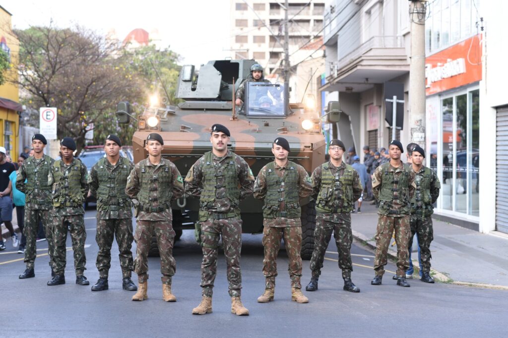 A imagem mostra um desfile de rua com muitas pessoas. No centro do desfile, há um grupo de pessoas carregando uma faixa com os dizeres "APAE PIRACICABA". Algumas pessoas estão em cadeiras de rodas, participando do desfile. Ao fundo, há edifícios com arquitetura clássica e moderna, e um céu parcialmente nublado. A rua está decorada com faixas e balões. A atmosfera geral parece ser de celebração e inclusão.
