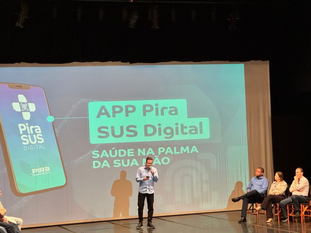 A imagem mostra uma apresentação em um palco. No centro, um homem está em pé, segurando um celular e um microfone, falando para a audiência. Atrás dele, um grande telão exibe o logotipo do "APP Pira SUS Digital" e a frase "SAÚDE NA PALMA DA SUA MÃO". À esquerda do telão, há uma representação de um smartphone com o mesmo logotipo do aplicativo. À direita, sentadas em cadeiras, estão três pessoas, possivelmente parte da audiência ou painelistas. O ambiente sugere um evento formal, como uma conferência ou lançamento de produto, com iluminação de palco e cortinas ao fundo.