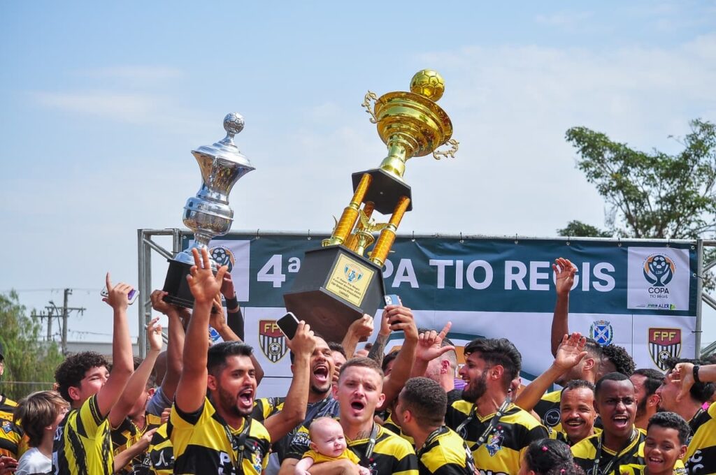 A imagem mostra um grupo de homens, alguns usando uniformes de futebol com listras amarelas e pretas, comemorando a vitória. Eles estão erguendo dois troféus: um prateado e um dourado, ambos com um futebol no topo. O troféu dourado é maior e mais ornamentado. Ao fundo, há uma faixa verde com letras brancas que diz "4ª COPA TIO REGIS". Há também logotipos de patrocinadores e a sigla "FPF". O céu está azul e claro, com algumas nuvens esparsas. A atmosfera geral é de celebração e alegria, com muitos dos homens sorrindo e gritando. Um bebê está sendo segurado por um dos homens no centro da imagem.