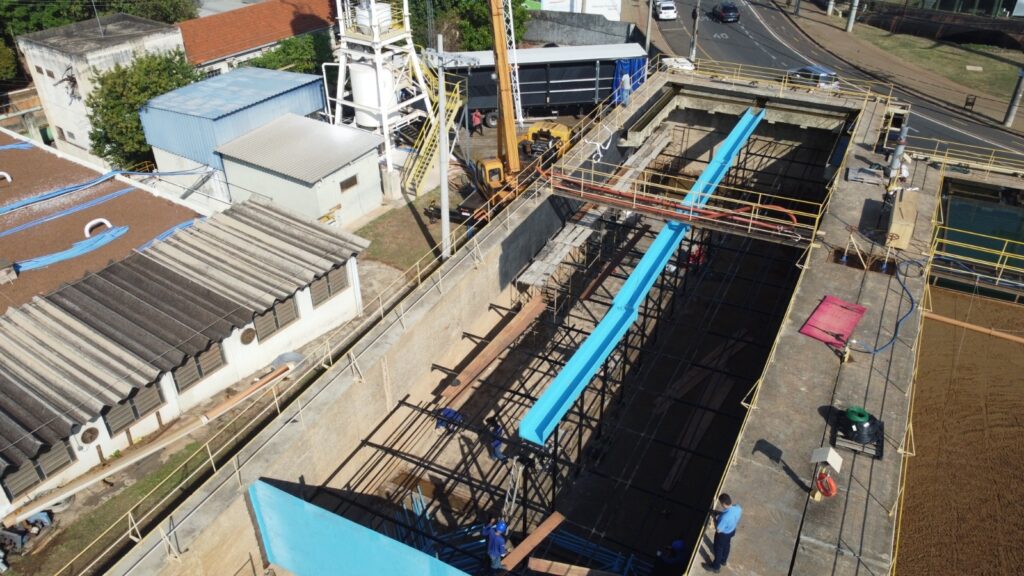 A imagem mostra uma vista aérea de uma estação de tratamento de água ou esgoto em construção ou em manutenção. * A área principal é um grande tanque retangular, com paredes de concreto, onde estão sendo instaladas estruturas metálicas e um canal azul. * Há trabalhadores no local, usando capacetes e uniformes, realizando atividades de construção. * Ao redor do tanque principal, há passagens elevadas com corrimãos amarelos, indicando acesso para manutenção e operação. * Em outras áreas da imagem, observam-se edifícios com telhados de amianto, tanques de armazenamento e tubulações. * Uma escavadeira amarela e um guindaste estão estacionados próximos a uma estrutura maior, possivelmente parte da infraestrutura da estação. * Ao fundo, uma rua com carros em movimento e vegetação indicam que a estação está localizada em uma área urbana ou semiurbana.