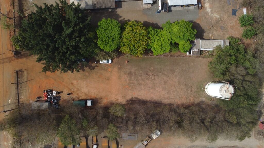 A imagem aérea mostra uma área com vegetação, construções e alguns veículos. No lado esquerdo, há uma grande árvore de folhagem densa e escura, com alguns carros estacionados próximos a ela. Abaixo da árvore, há um amontoado de materiais, incluindo pneus e tubos, e um pequeno galpão. Um caminhão verde também está visível na área. A parte central da imagem é dominada por um terreno de terra batida, com algumas árvores de folhagem verde vibrante ao longo da borda superior. Uma construção com telhado cinza está situada entre as árvores. No lado direito, há um grande tanque branco cilíndrico, cercado por árvores e vegetação. Mais ao fundo, vê-se uma construção com telhado branco e algumas estruturas menores. A paisagem geral sugere uma área rural ou industrial com presença de natureza.