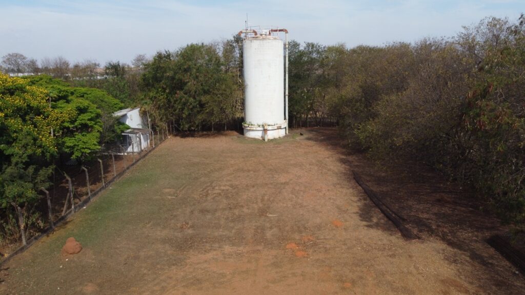 A imagem mostra um grande reservatório de água branco, de formato cilíndrico, cercado por árvores. Há uma pequena construção ao lado esquerdo do reservatório, com uma cerca de arame ao redor. O terreno em primeiro plano é de terra batida e grama, com algumas áreas de vegetação seca. O céu está azul e claro.