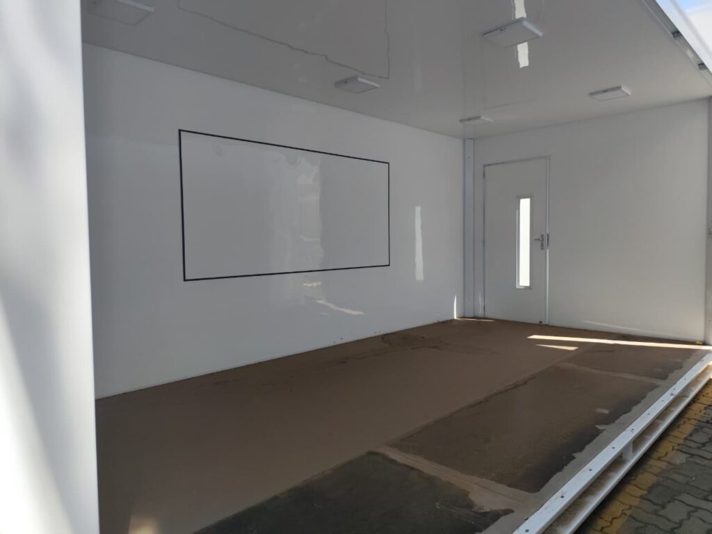 A imagem mostra o interior de um espaço branco e limpo, possivelmente um trailer ou um contêiner. Na parede esquerda, há um grande retângulo preto delineado, que pode ser uma tela ou um quadro branco. O piso é de cor marrom e parece ser feito de um material resistente, possivelmente cimento ou concreto. Na parede do fundo, há uma porta branca com uma janela vertical estreita e uma maçaneta prateada. O teto é branco e possui várias luminárias embutidas. A iluminação é clara e uniforme, destacando a limpeza e a organização do espaço.