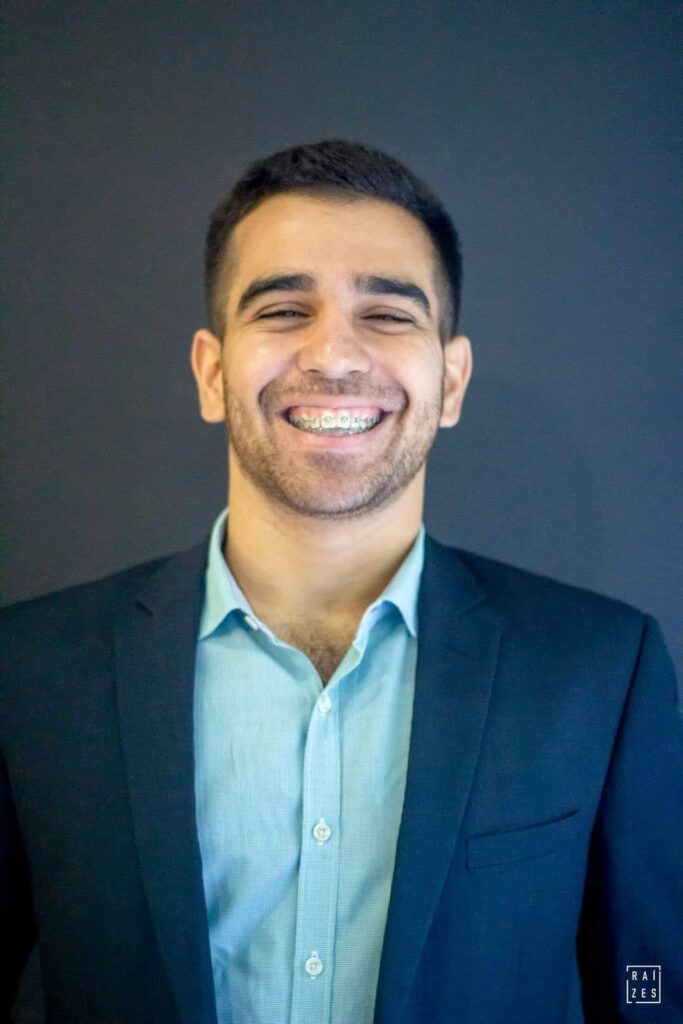 A imagem mostra um homem jovem sorrindo, vestindo um terno escuro e uma camisa azul clara. Ele tem aparelho nos dentes, que são visíveis em seu sorriso aberto. O fundo é um tom escuro e sólido, possivelmente um estúdio. A iluminação destaca o rosto do homem e a textura de suas roupas. Um pequeno logotipo com as letras "RAI ZES" aparece no canto inferior direito da imagem.