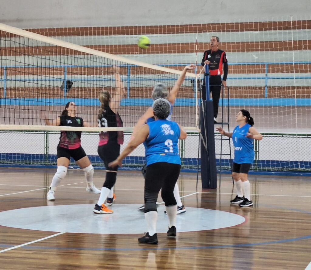 A imagem mostra uma partida de vôlei em uma quadra interna. Há seis jogadoras em quadra, divididas entre os dois lados da rede. Uma jogadora está no ar, prestes a sacar ou atacar a bola, que está em voo acima da rede. Um árbitro está posicionado em uma cadeira alta ao lado da rede, observando a partida. As jogadoras usam uniformes de cores diferentes, alguns azuis e outros pretos com detalhes em rosa e verde. A quadra tem piso de madeira e as paredes são decoradas com listras azuis e marrons.